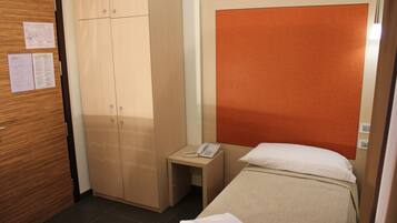 A hotel room with a bed, a wooden wardrobe, a small bedside table with a phone, and a wall-mounted bulletin board.