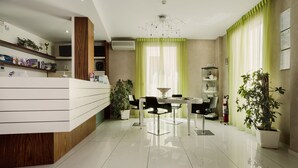 Un área de recepción moderna con un mostrador blanco, una lámpara de araña y cortinas verdes.
