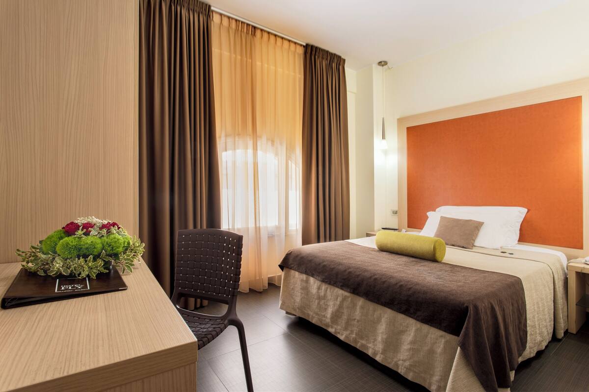 A hotel room with a bed, a desk, and a chair. There is a window with curtains, a nightstand, and a lamp.