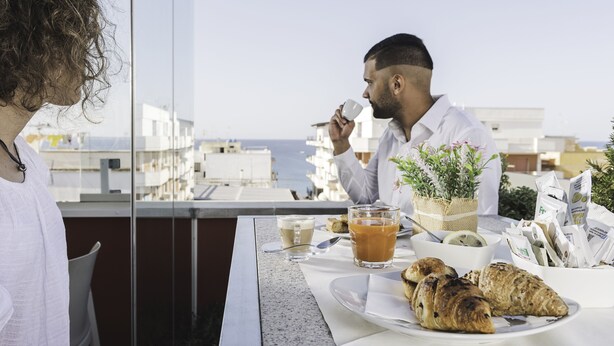 Desayuno con cruasanes, café y jugo sobre una mesa con vista al mar y edificios.