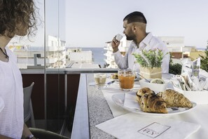 A breakfast setting with croissants, coffee, and juice on a table with a view of the sea and buildings. - Hotel 33 Baroni (Gallipoli)