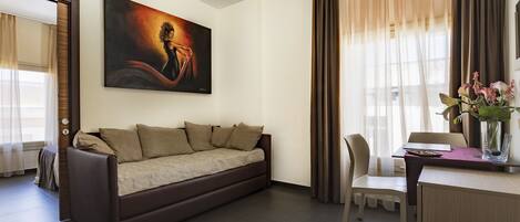 A modern hotel room with a brown sofa, a dining table with chairs, a painting on the wall, and a window with curtains.