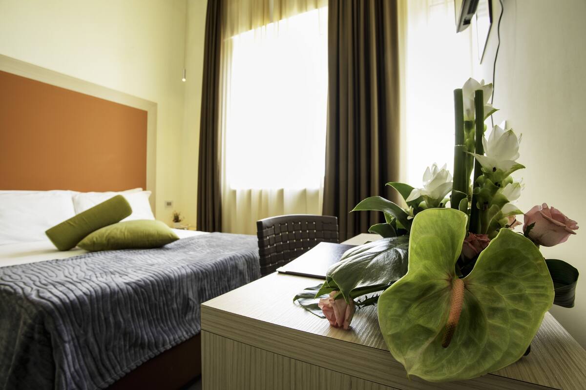 A hotel room with a bed, a desk with a laptop, and a large plant with flowers.