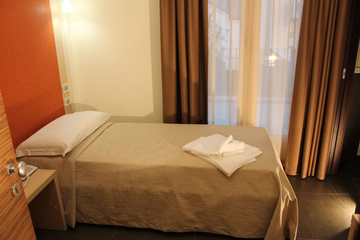 A hotel room with a bed, a nightstand, a lamp, and a window with curtains.