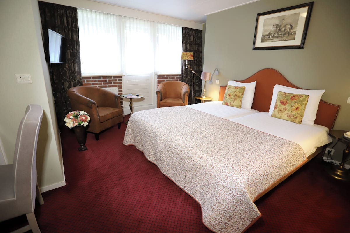 Une chambre d’hôtel avec un grand lit, un coin salon comprenant un fauteuil et une petite table, un téléviseur fixé au mur et un tableau encadré au-dessus du lit.
