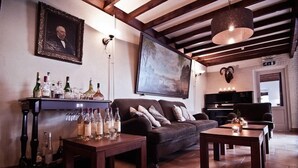 Een gezellige woonkamer met een donkerbruine bank, een houten salontafel en een open haard. Op een plank staan wijnflessen en glazen, en aan de muur hangt een groot schilderij.