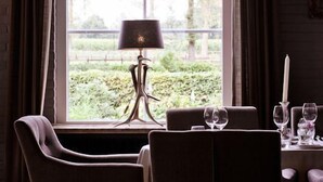 Een eetruimte met een tafel voor twee, een lamp en uitzicht op groen buiten een raam.