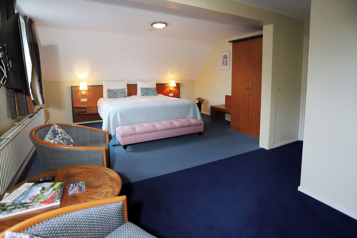 Une chambre d’hôtel avec un lit, un banc rose, une table d’appoint en bois, une chaise et un téléviseur.