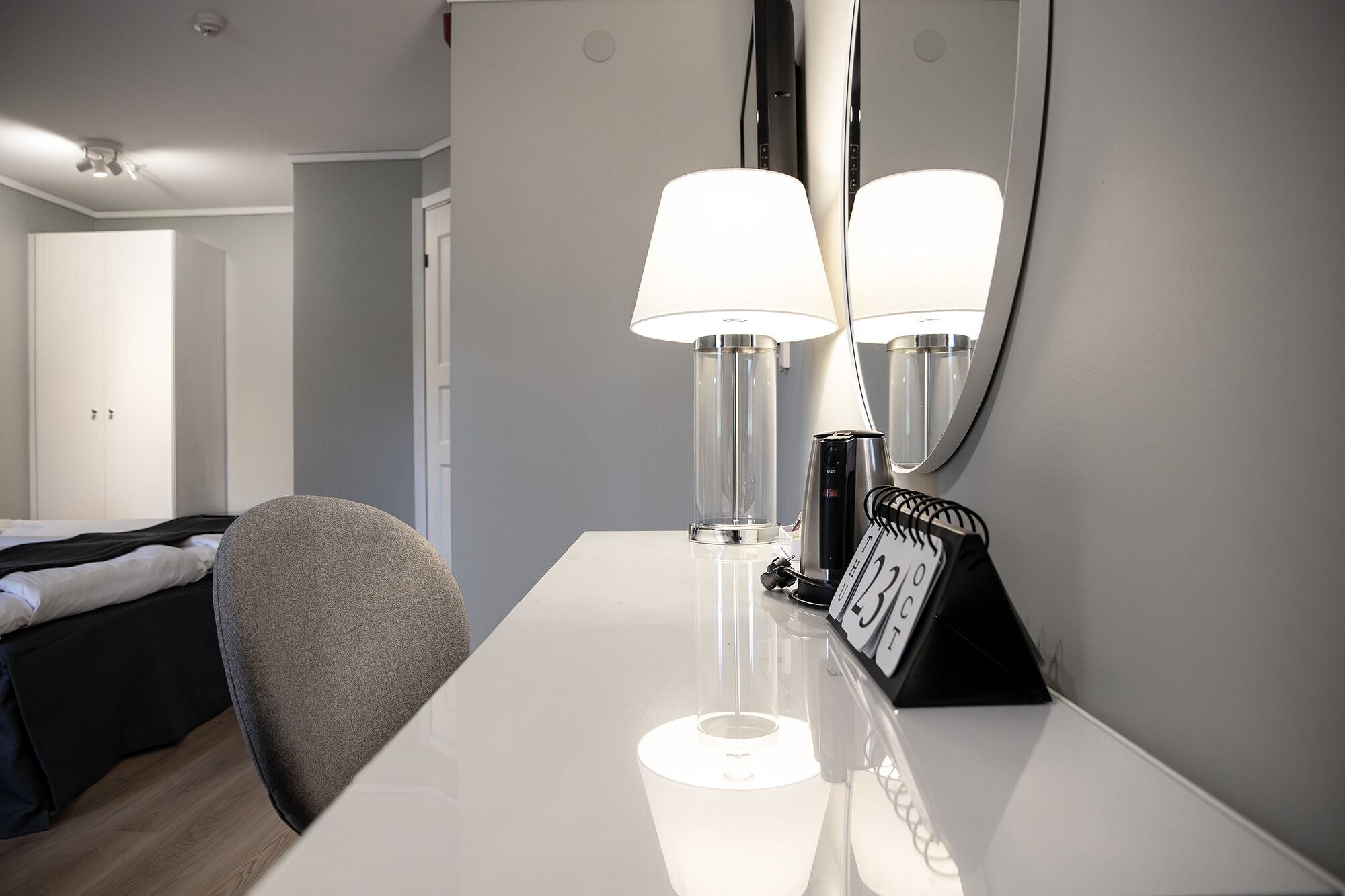 A hotel room with a bed, a desk with a lamp, a mirror, and a calendar.