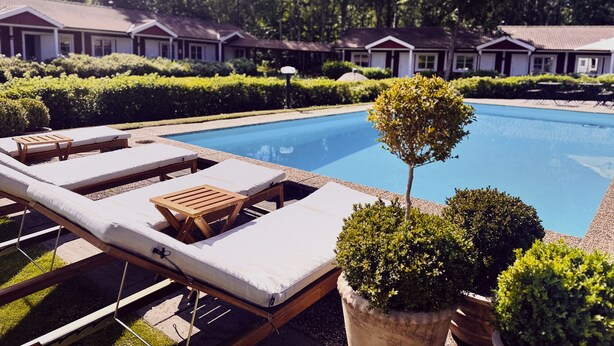 Una piscina con sillas de descanso y plantas en macetas, rodeada de setos cuidados y una hilera de casas al fondo.