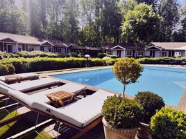 Piscina com espreguiçadeiras e plantas em vasos, rodeada por sebes bem aparadas e uma fileira de casas ao fundo.