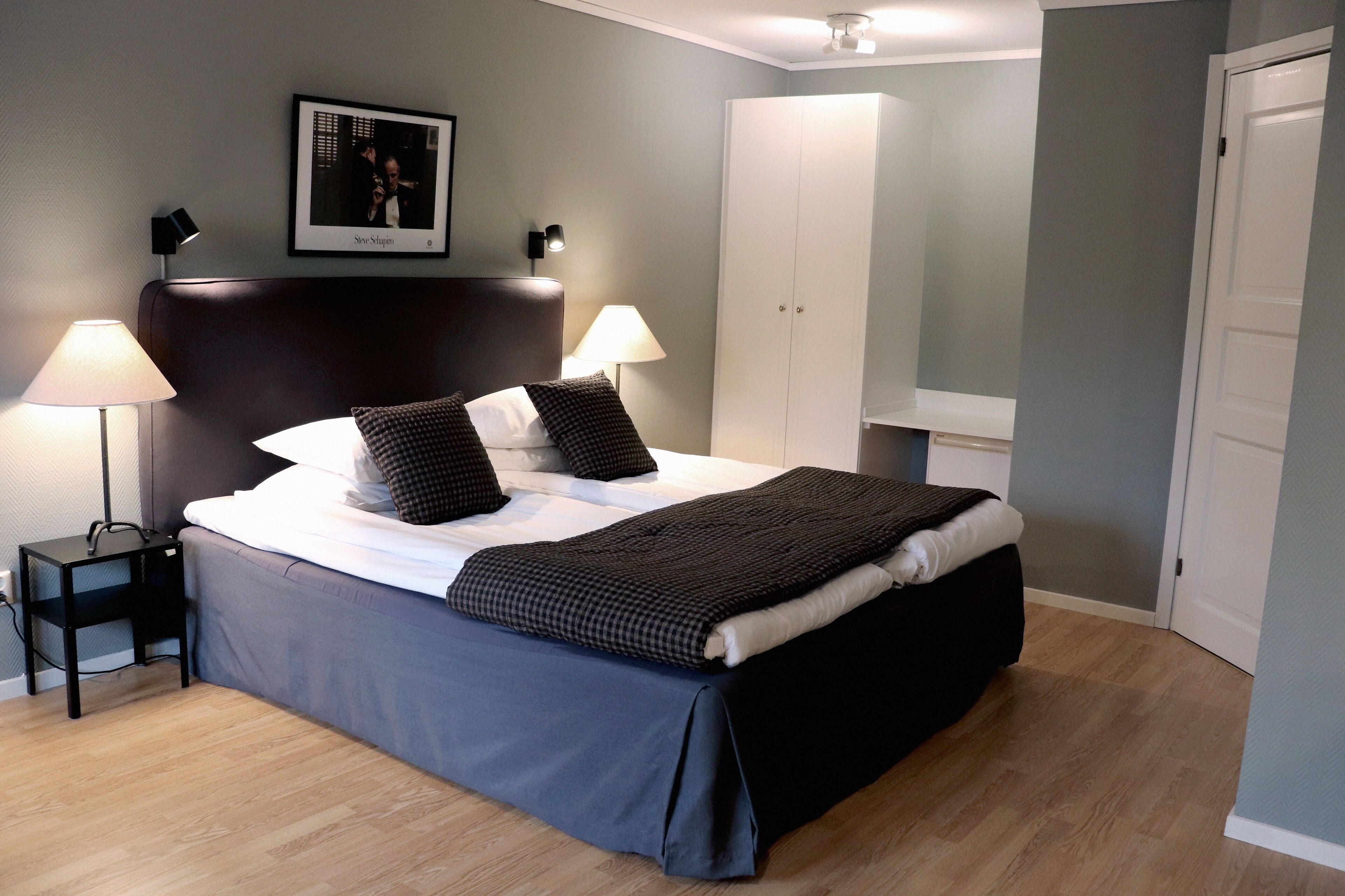 Cama bem arrumada com cabeceira escura, duas luminárias de apoio, um quadro na parede e um guarda-roupa ao fundo.