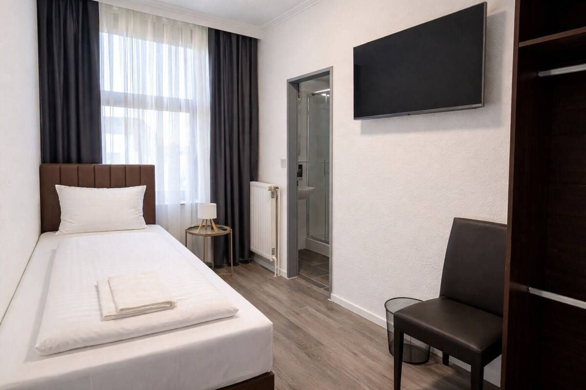 A hotel room with a bed, a chair, a TV, a window with curtains, and a door leading to a bathroom.