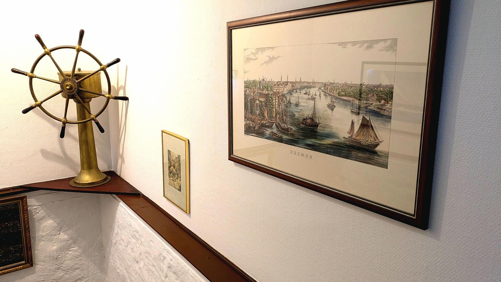 Eine gerahmte historische Illustration einer Hafenszene, ein Messing-Steuerrad und ein kleines gerahmtes Bild an einer Wand.