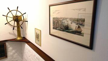 Eine gerahmte historische Illustration einer Hafenszene, ein Messing-Steuerrad und ein kleines gerahmtes Bild an einer Wand.