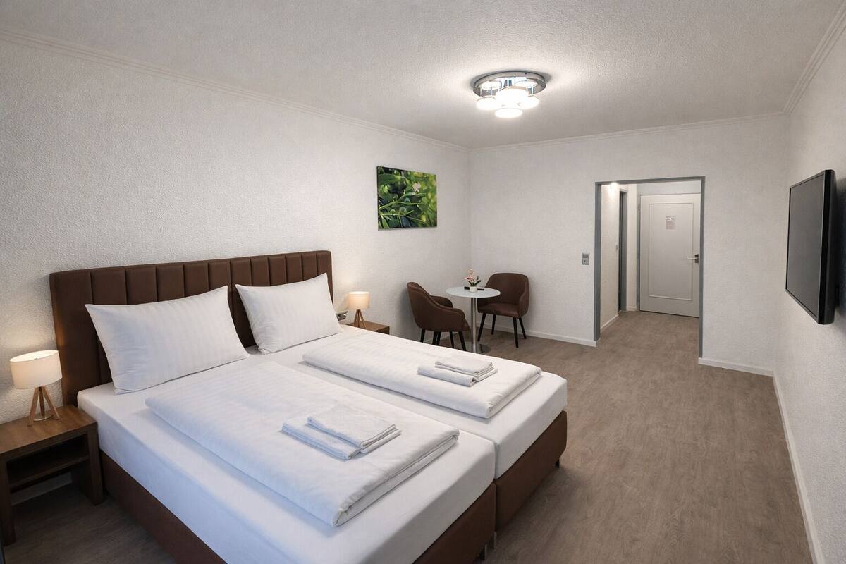 A hotel room with a double bed, two bedside tables with lamps, a small round table with chairs, a television mounted on the wall, and a door leading to another room.