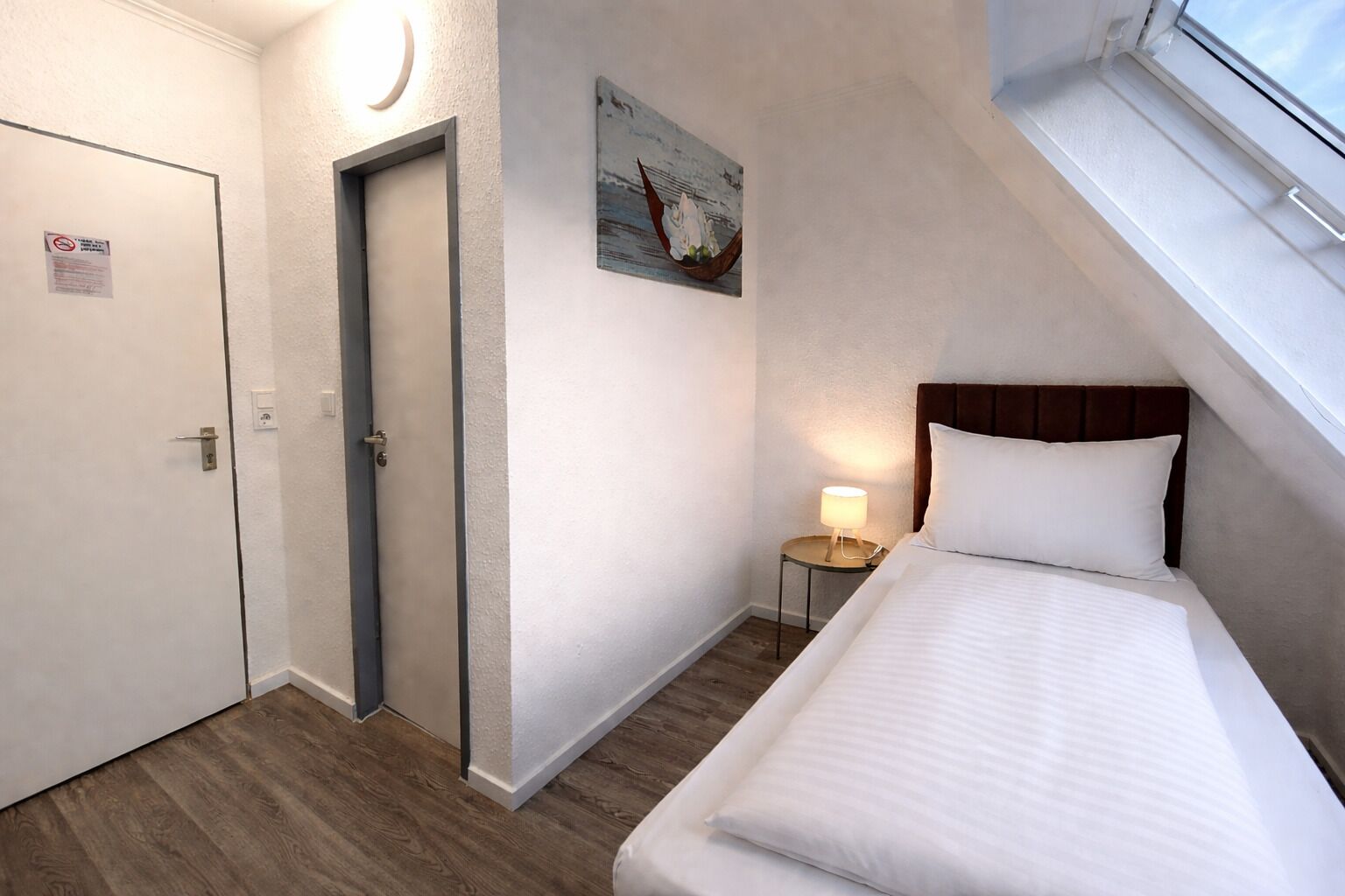 Ein Hotelzimmer mit Bett, Nachttisch mit Lampe, Tür und einem Gemälde an der Wand.
