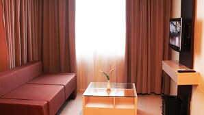 A living room with a sofa, a glass coffee table with a vase, a TV on a stand, and curtains. - Solo Paragon Hotel & Residences (Surakarta)