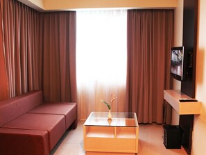 A living room with a sofa, a glass coffee table with a vase, a TV on a stand, and curtains. - Solo Paragon Hotel & Residences (Surakarta)