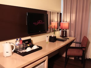 A hotel room with a TV, a minibar, a coffee maker, a tray with cups and a bottle, a remote control, and a chair. - Solo Paragon Hotel & Residences (Surakarta)