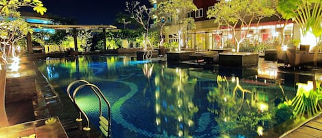 Una piscina iluminada de noche con agua cristalina, rodeada de zonas de descanso bien iluminadas y un edificio de varios pisos con balcones.