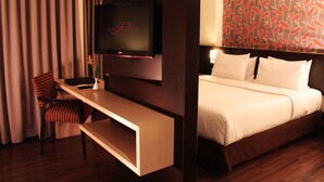 A hotel room with a bed, a TV mounted on the wall, a desk with a chair, and a patterned wall. - Solo Paragon Hotel & Residences (Surakarta)