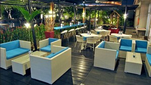 An outdoor seating area with wicker furniture and tables, surrounded by greenery and a covered structure. - Solo Paragon Hotel & Residences (Surakarta)