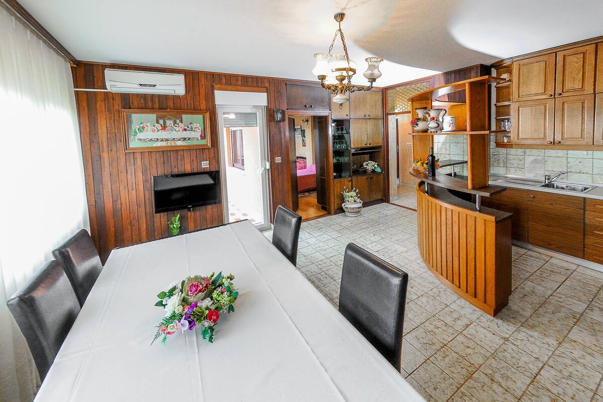 Comedor con una mesa blanca, sillas y un centro de mesa floral. Se observa una cocina con armarios de madera y un lavamanos.