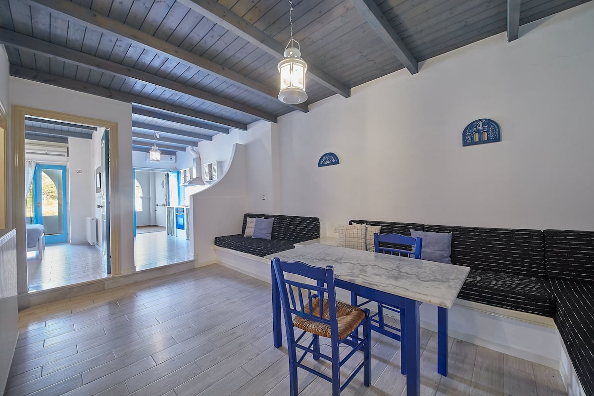 A spacious room with a wooden ceiling, a dining table with blue chairs, a black sofa with cushions, and a hanging light fixture.