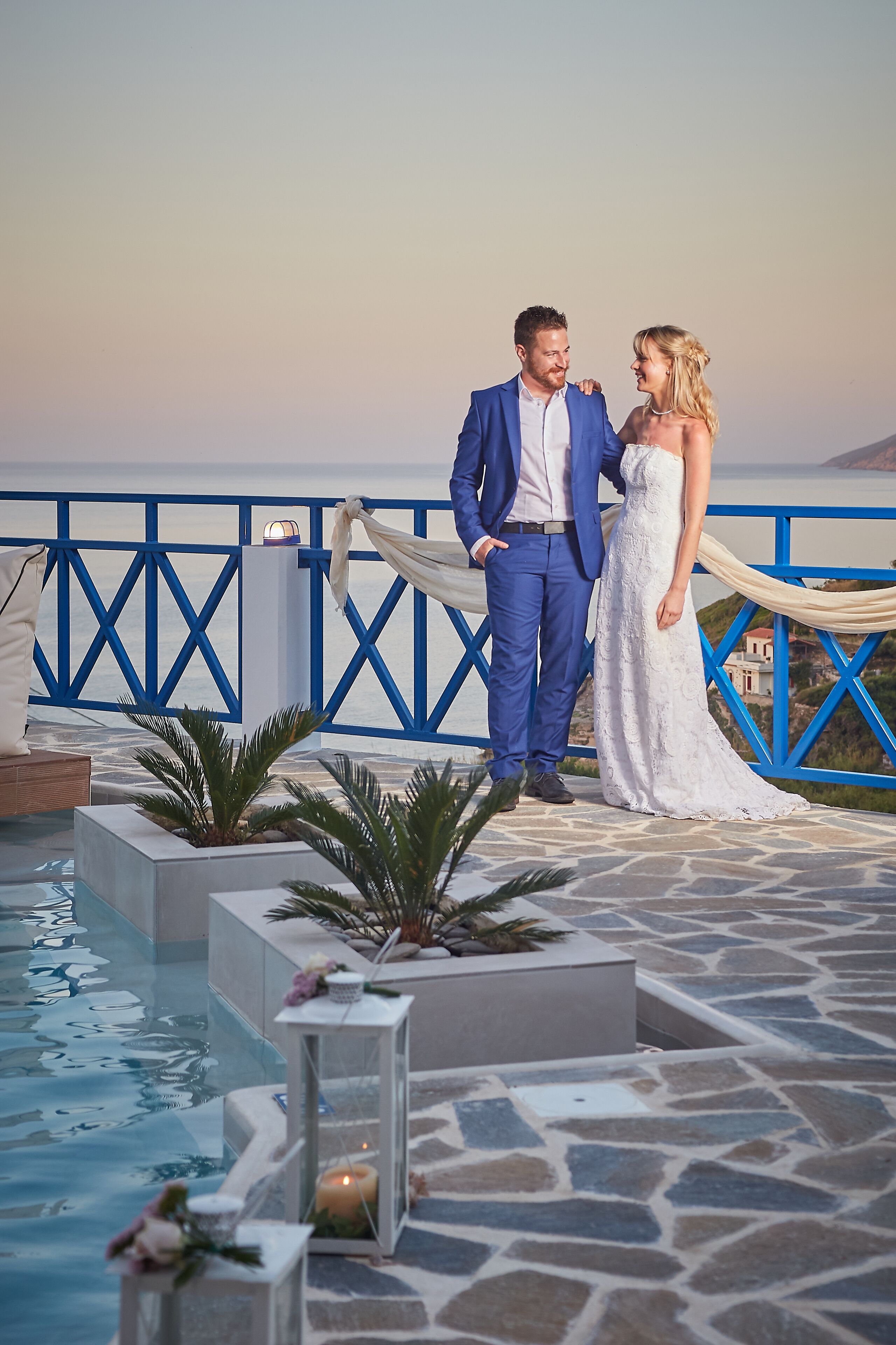 A couple standing by a pool with a blue railing and a view of the sea.
