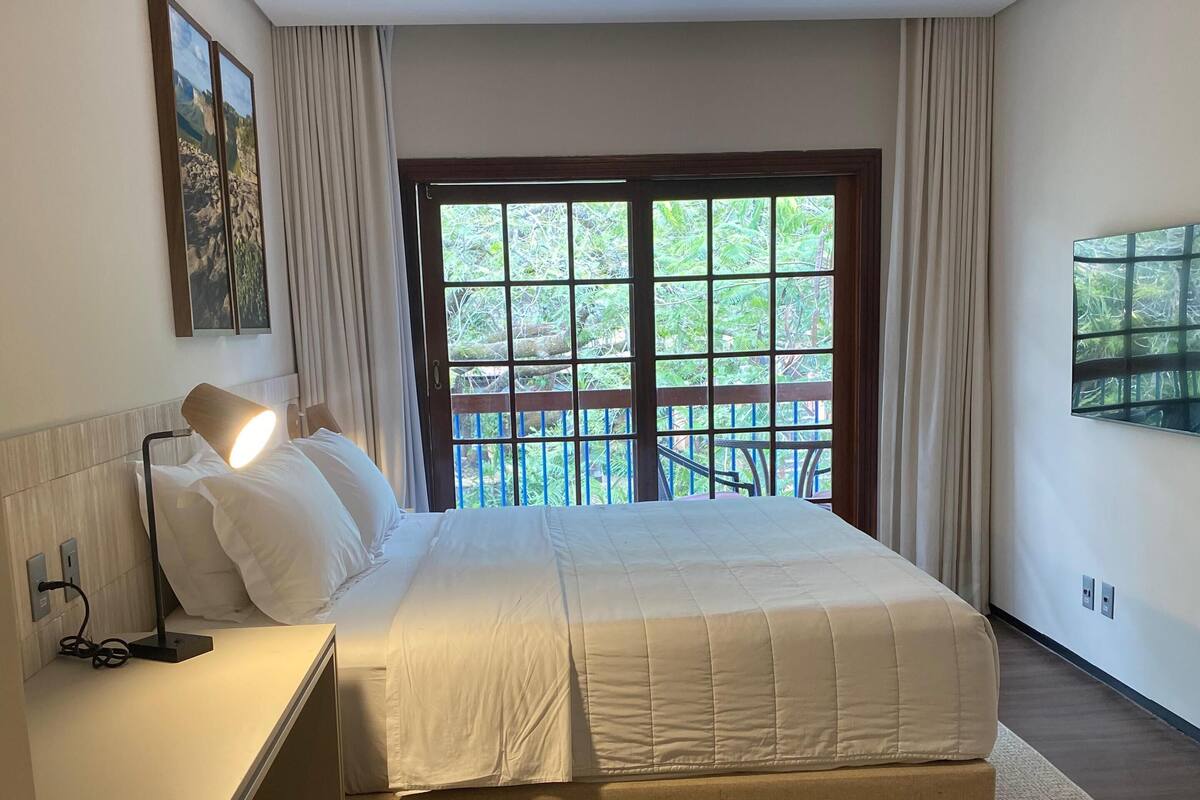 Un dormitorio moderno con una cama grande, una mesita de noche con lámpara, vista a zonas verdes a través de una ventana, y un plafón.