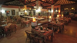 A restaurant with multiple dining tables and chairs, a central bar area, and a high ceiling with exposed beams.