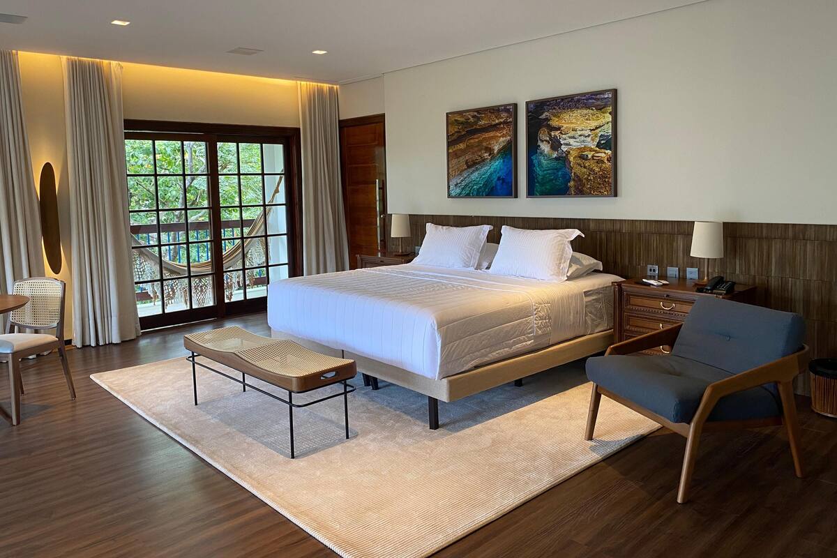 Un hotel moderno con una cama grande, un área de descanso con una silla, una mesita y vistas a la vegetación por la ventana.