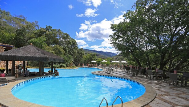 A swimming pool with a tiled deck, lounge chairs, umbrellas, and a thatched-roof pavilion.