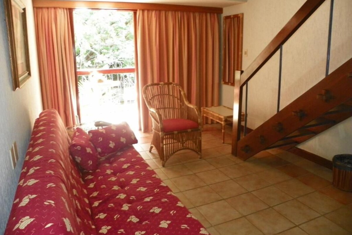A living room with a sofa, a wicker chair, a staircase, and a view through a window with curtains.