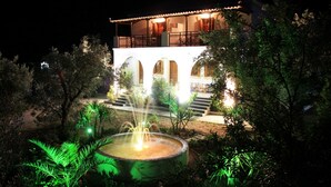 Un edificio de dos pisos con balcón, una fuente y un jardín bien cuidado de noche.