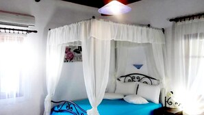 Una habitación con una cama bajo un dosel blanco, una colcha azul, un aire acondicionado de pared y una mesita con una lámpara.
