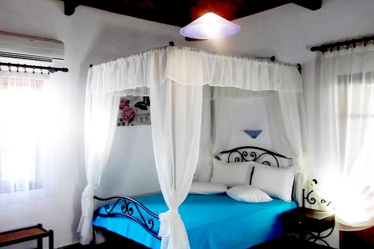 A room with a bed under a white canopy, a blue bedspread, a wall-mounted air conditioner, and a small table with a lamp.