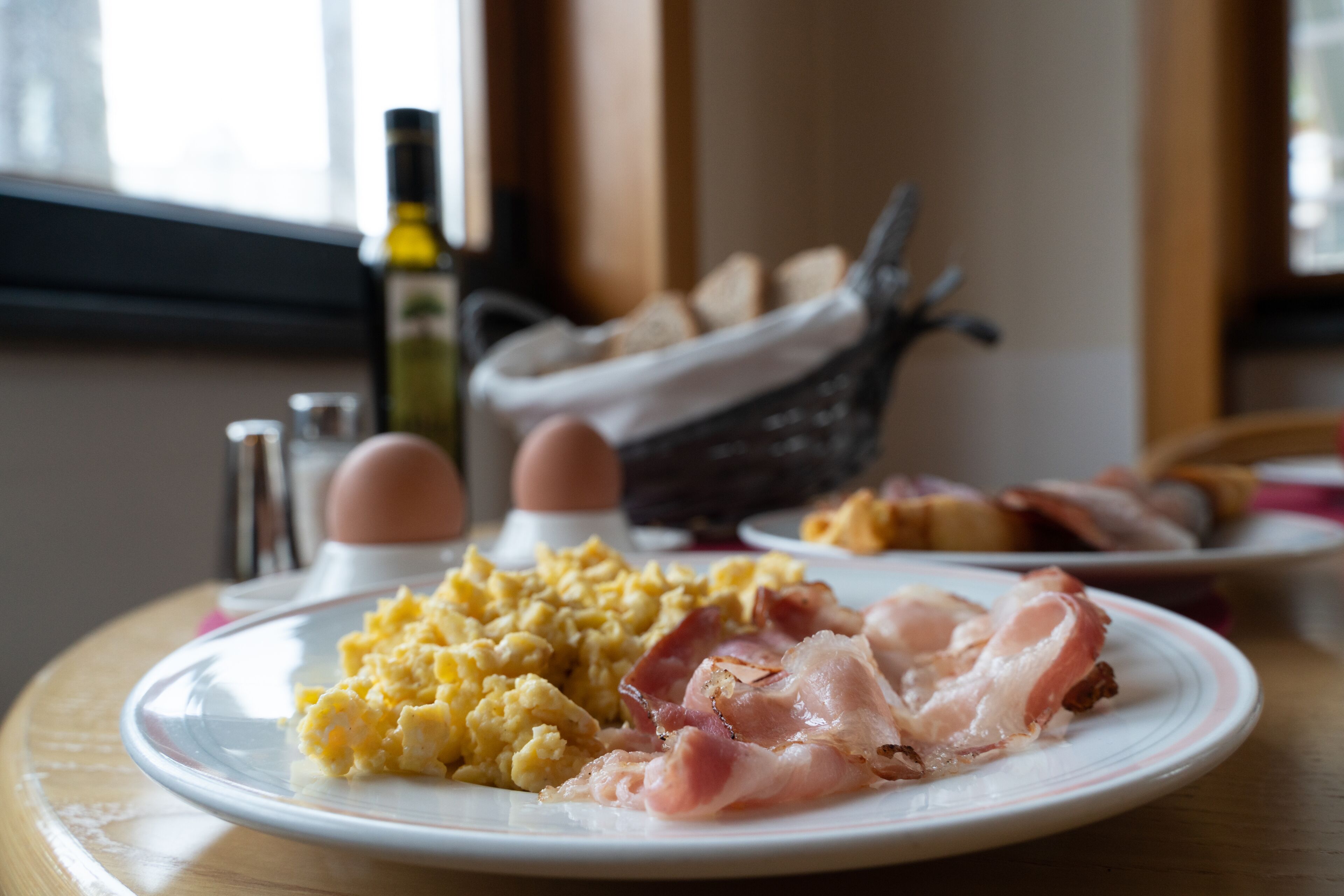 Un plato de desayuno con huevos revueltos y tocino, un salero y una botella de aceite de oliva sobre una mesa de madera.