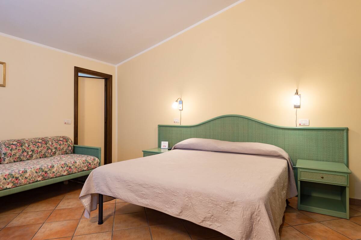 A bedroom with a bed, a nightstand, a green headboard, a floral sofa, and a wall-mounted light fixture.