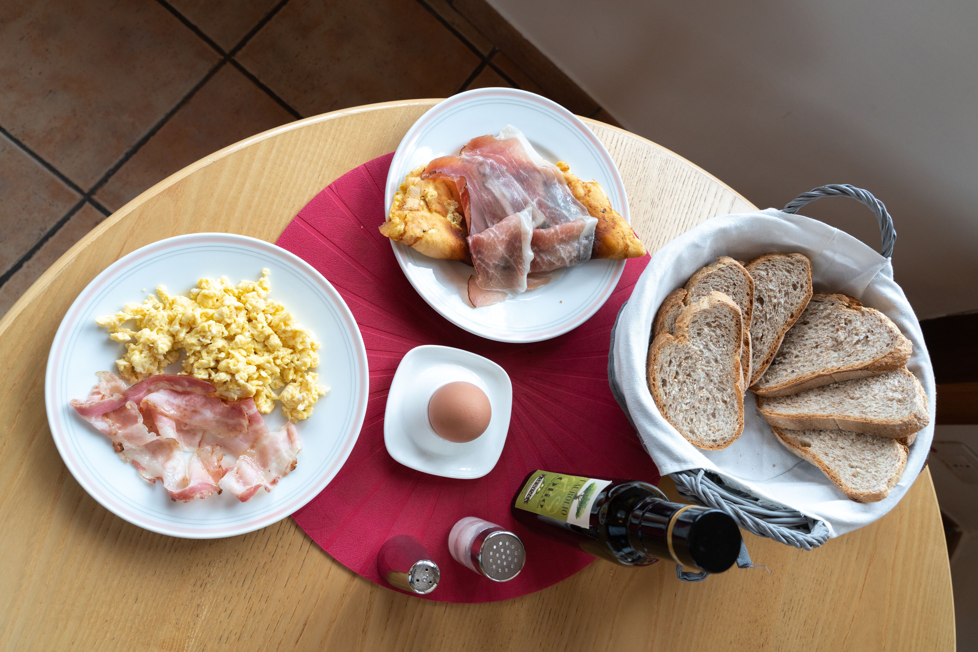 Una mesa de desayuno con platos de huevos revueltos, tocino, un croissant y tostadas, junto con salsas y una botella de aceite de oliva.