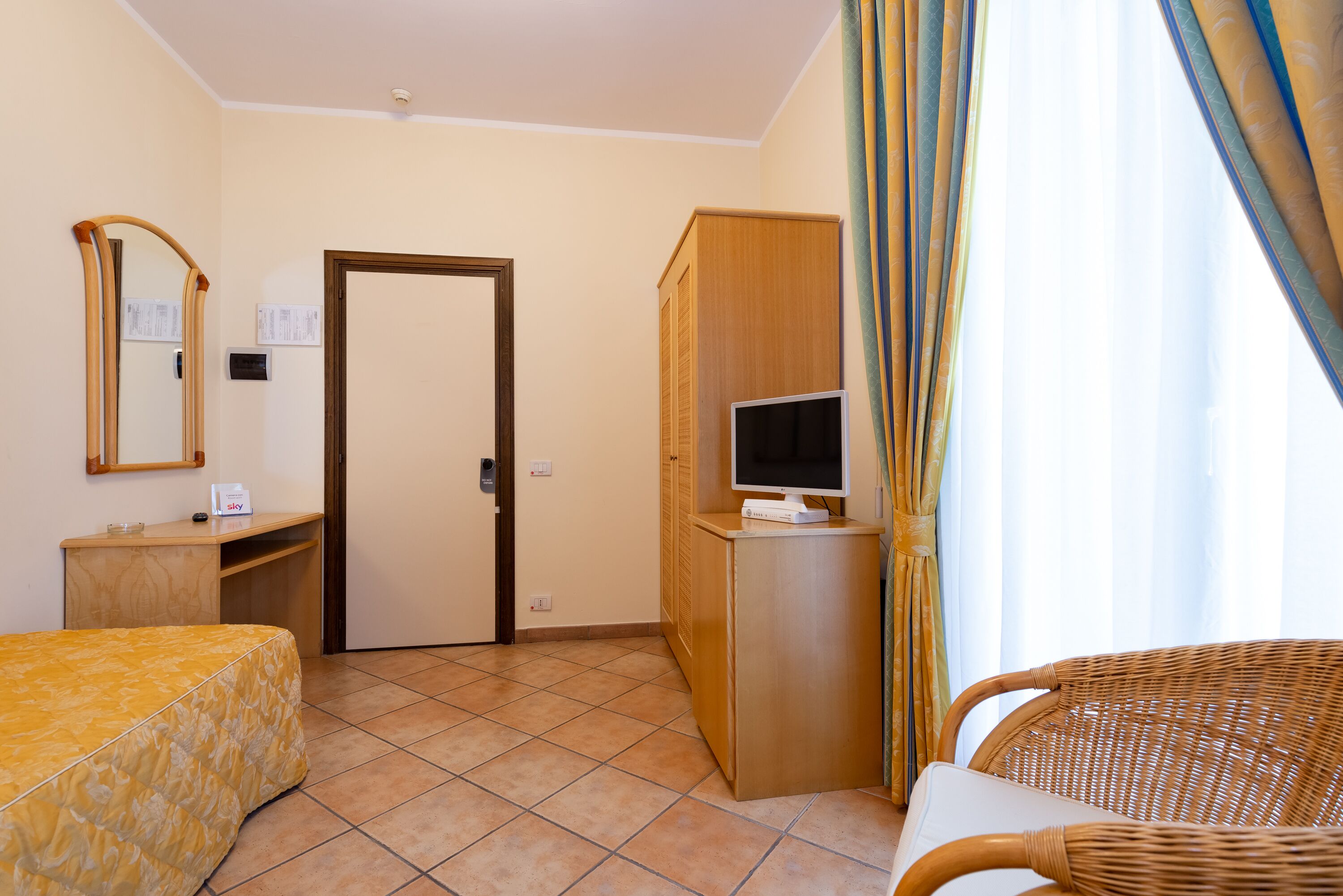 A hotel room with a bed, a desk, a television on a cabinet, a wicker chair, and a door.