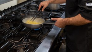A chef cooking on a commercial stove in a kitchen with stainless steel appliances and a tiled backsplash.