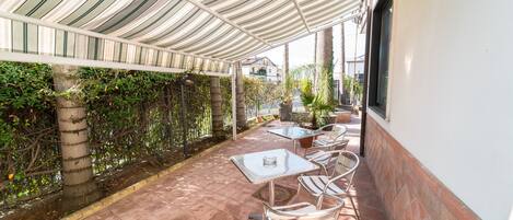 An outdoor patio with tiled flooring, metal chairs, and tables under a striped canopy.