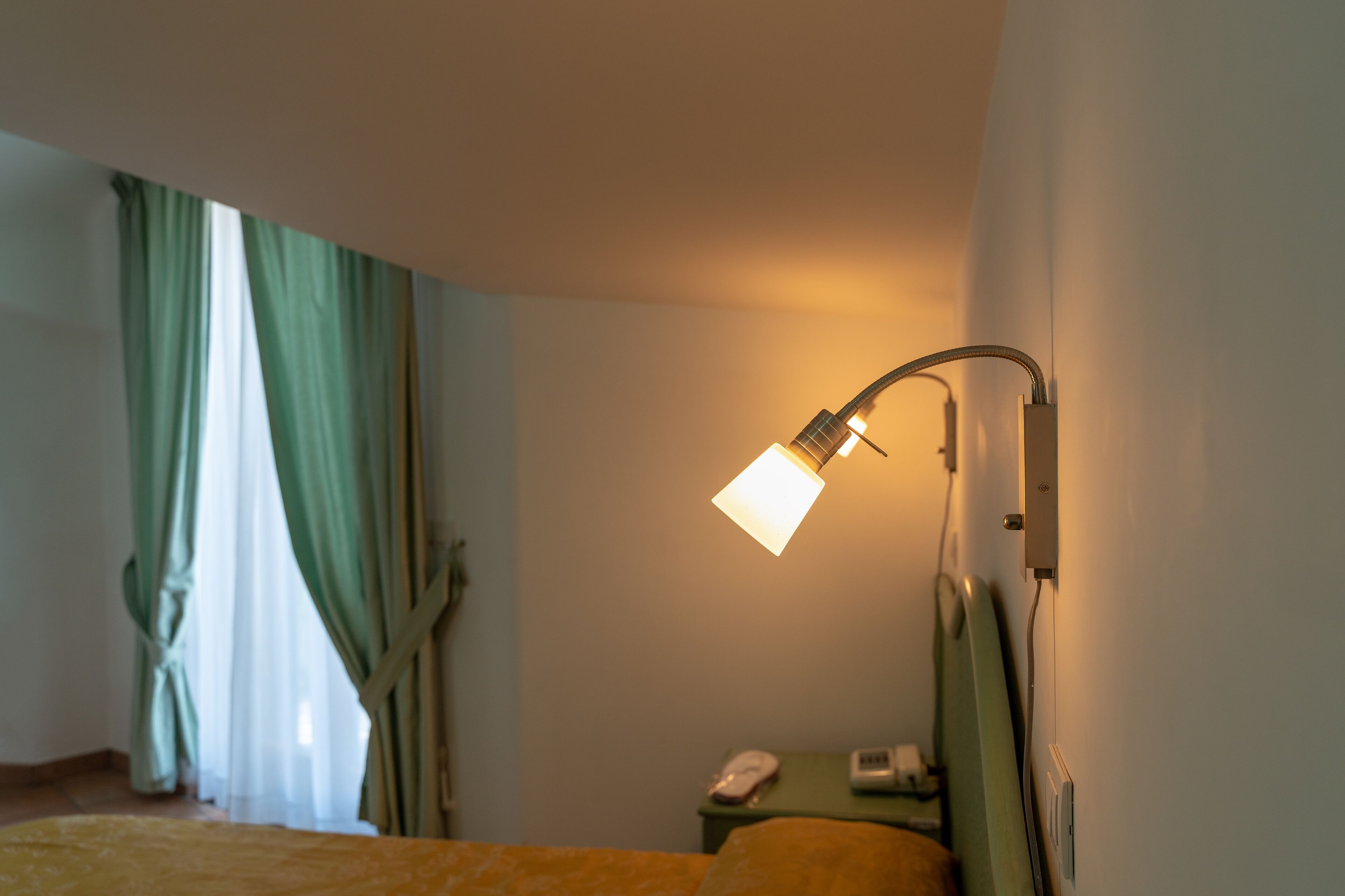 A hotel room with a bed, a bedside table with a lamp, and a window with curtains.
