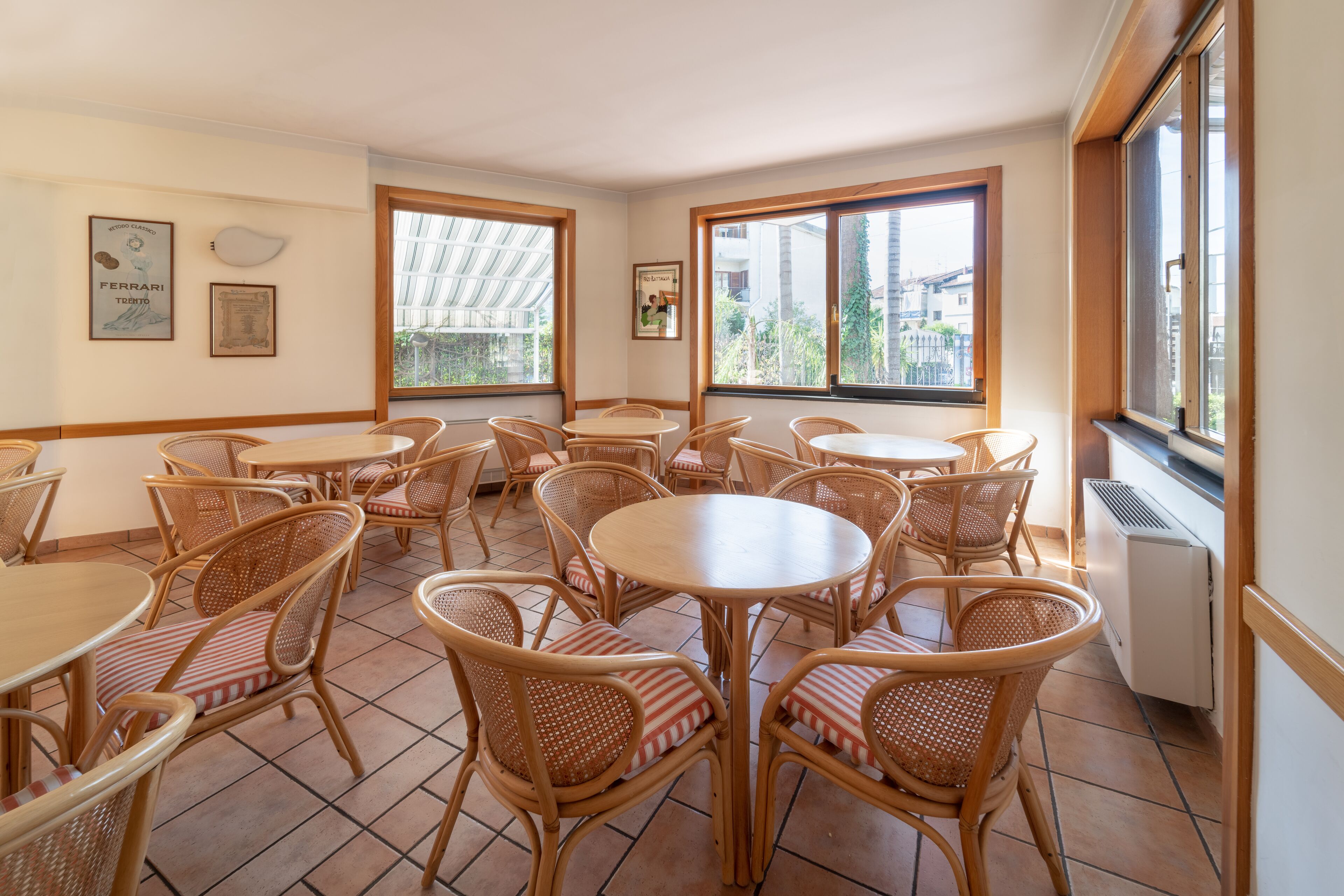 A dining area with multiple round tables and wicker chairs, large windows with views of buildings, and a tiled floor.
