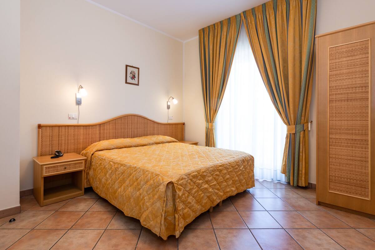 A hotel room with a large bed, bedside lamps, a nightstand, a wardrobe, and a window with curtains.