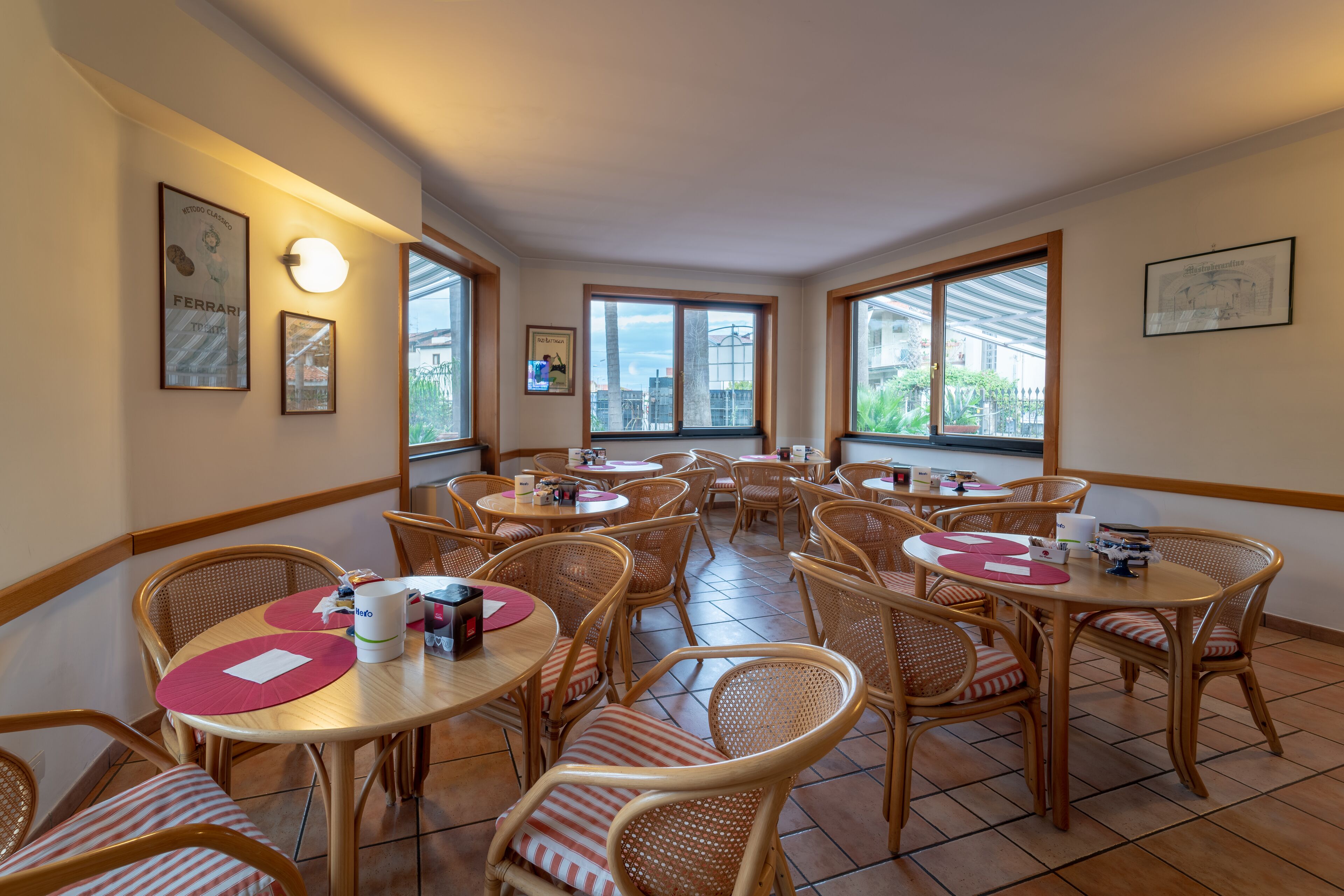A dining area with multiple tables and wicker chairs, featuring framed artwork on the walls and large windows providing natural light.