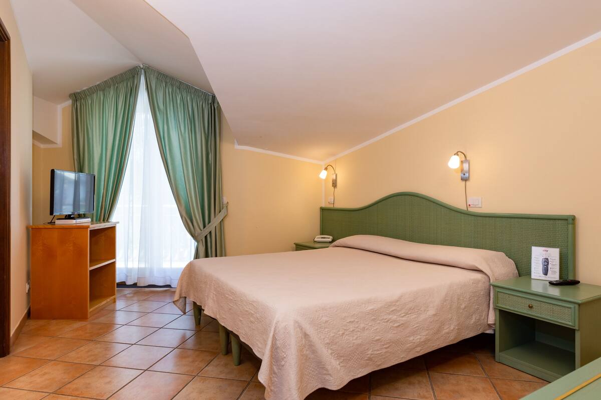 A hotel room with a bed, a nightstand, a TV on a wooden stand, and a window with curtains.