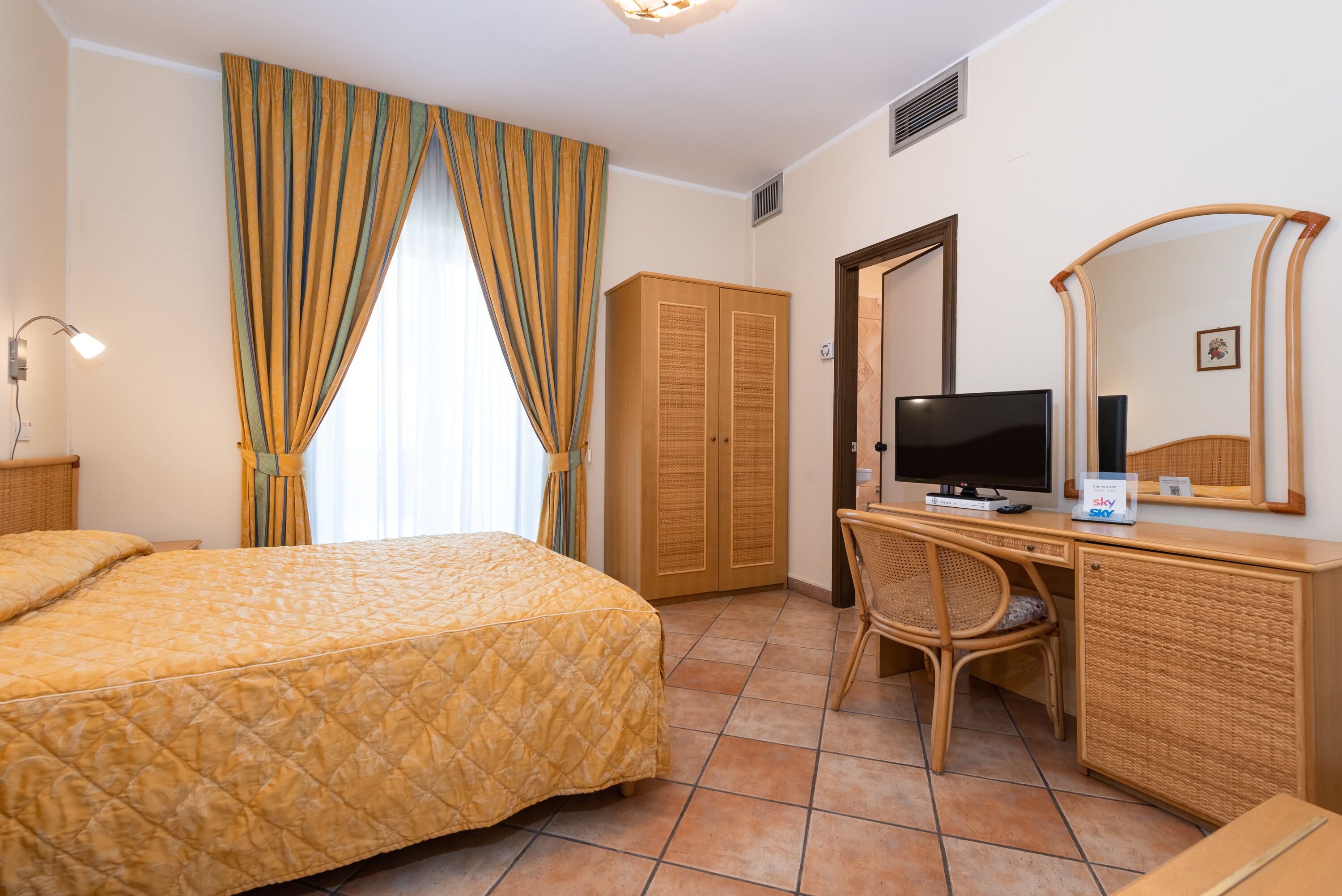 Habitació d'hotel amb un llit, un escriptori amb televisió, una cadira, un armari i un mirall gran.